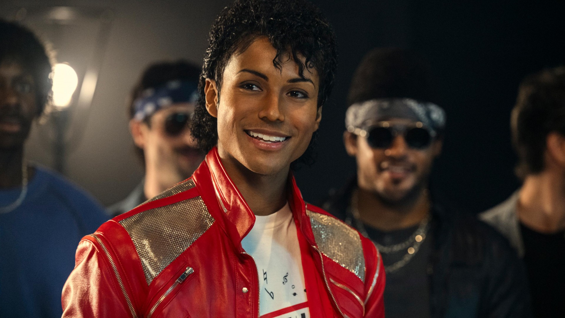 Smiling young man in a red metallic jacket in the foreground, with friends blurred in the background at a social event.