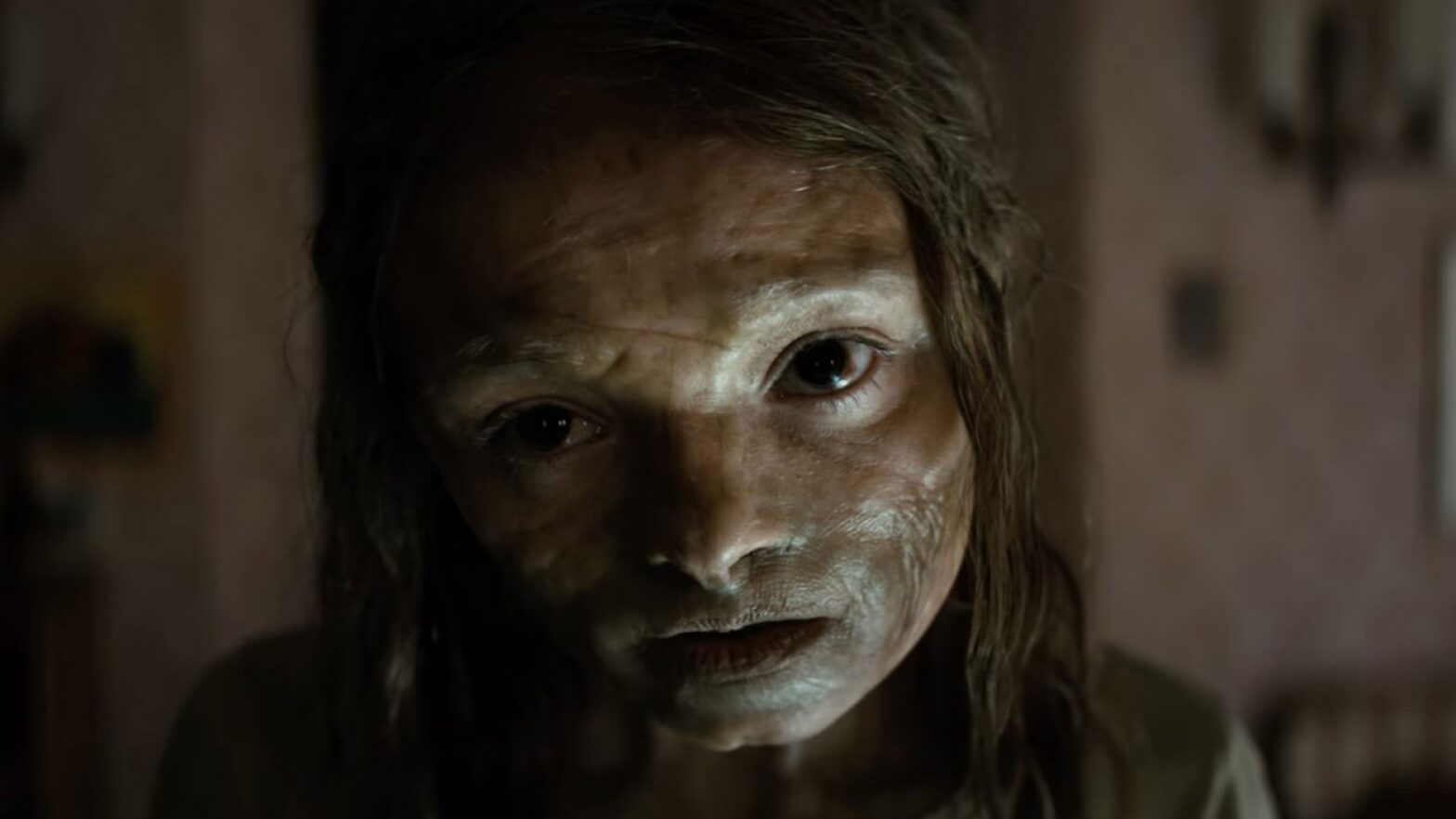 Close-up of a person with pale, smeared makeup in a dimly lit room, staring at the camera.