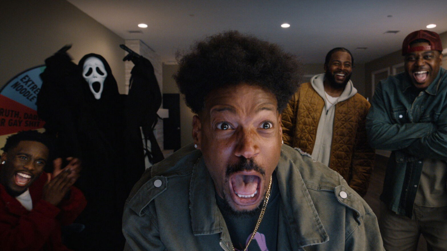 Man in the foreground screams with mouth wide open as friends laugh indoors; a ghost-costume figure is visible in the back left.