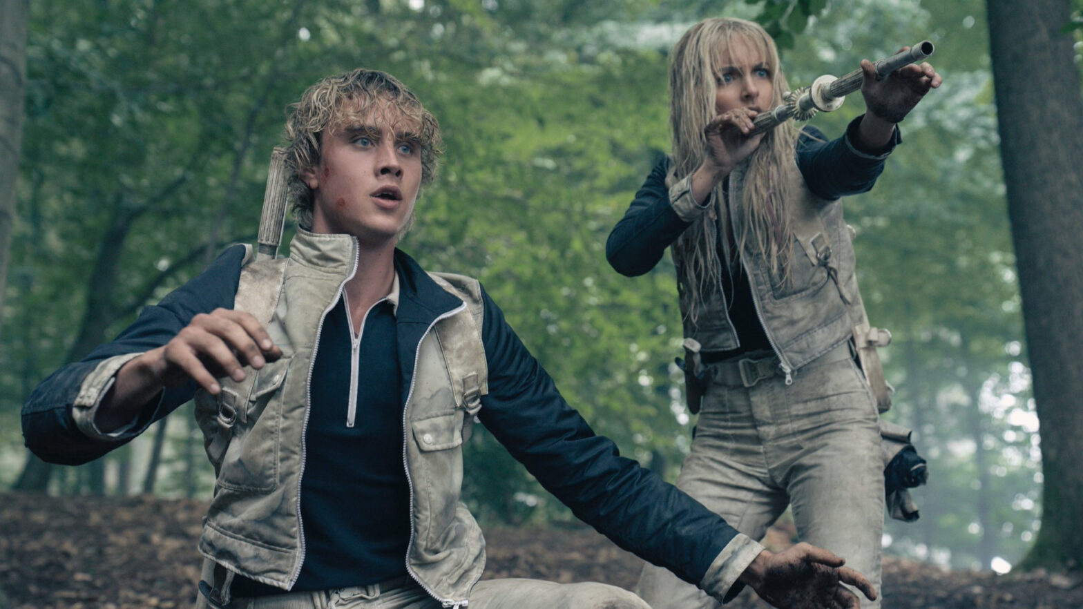 Two young survivors in beige vests crouch in a forest, one with a spear-like weapon raised, both tense and alert.
